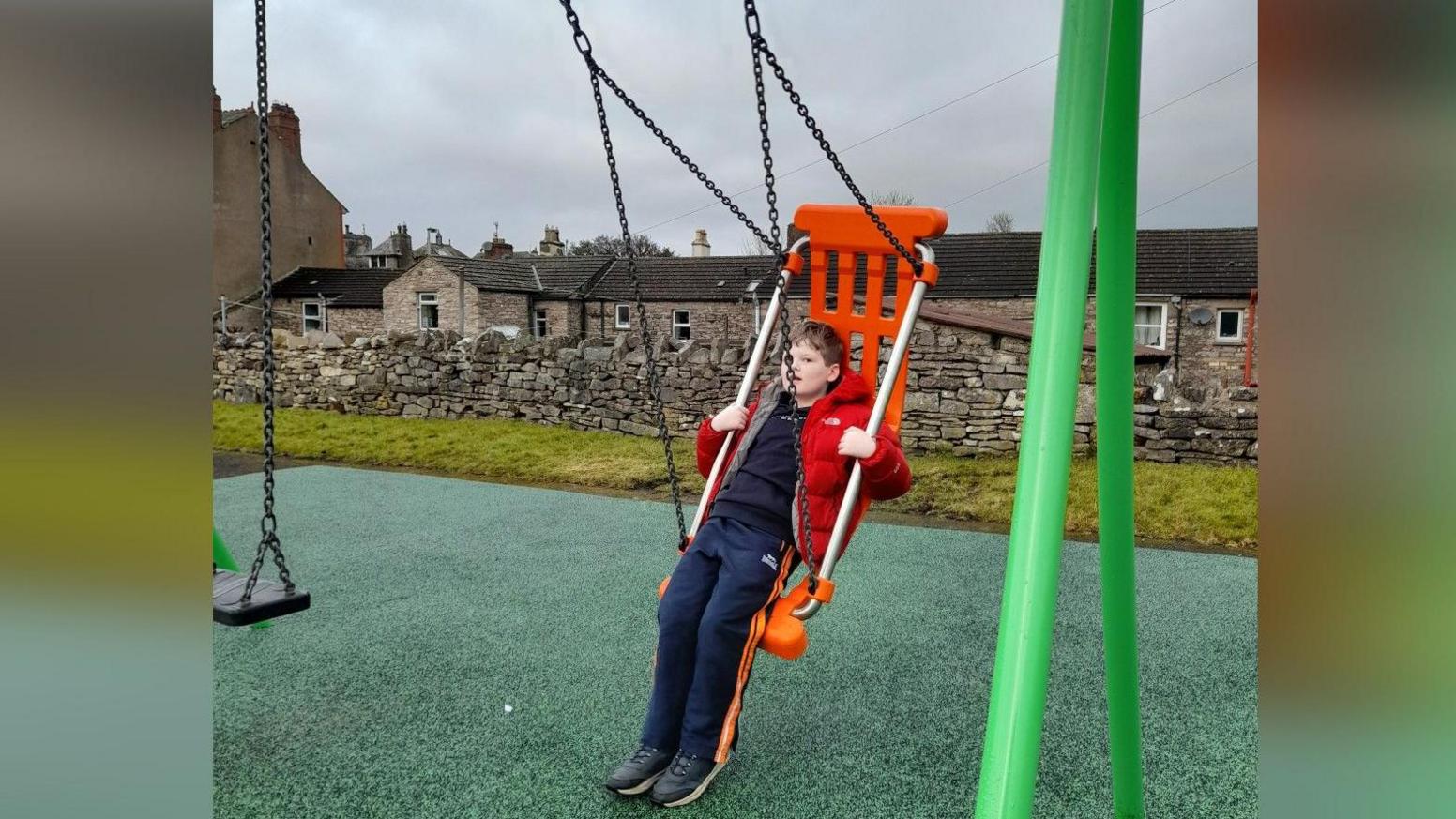 Alex on a large adapted swing in a playground. It has a green frame and orange seat. He is leaning on the seat and looking at the camera. He is wearing a navy blue tracksuit and red coat. He has short brown hair. Behind him is a drystone wall and some stone cottages.