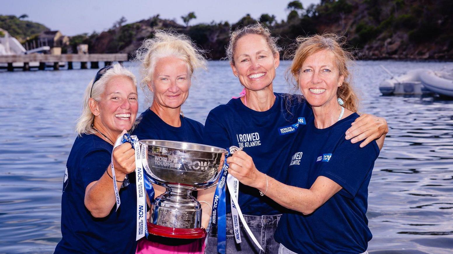 Four women holding a silver trophy that reads "World's Toughest Row". They are standing in front of the sea and are all wearing the same navy top. They are all smiling into the camera. 