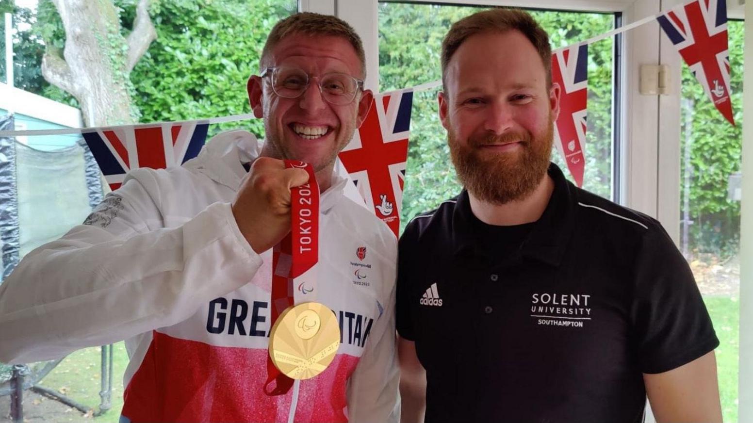 A picture of Aaron Phipps in a Great Britain tracksuit holding his gold medal and Jon Cooper standing in front of Union Jacks in front of a garden. 