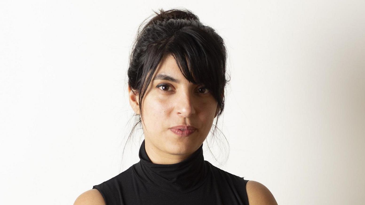 Yasmin poses in sleeveless black high-neck top. She has long black hair worn up and a fringe and dark eyes.