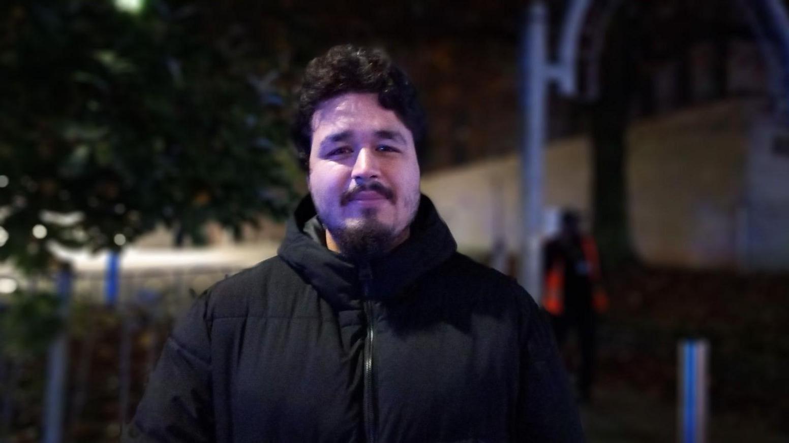 Ali Daniel facing the camera wearing a black coat at night. He has dark hair and a moustache.