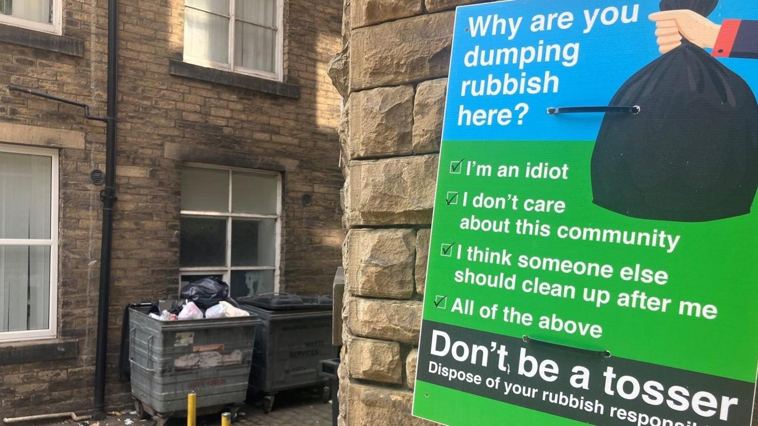 The entrance to a Bradford passageway with several large bins in it and a large green sign urging people not to dump rubbish