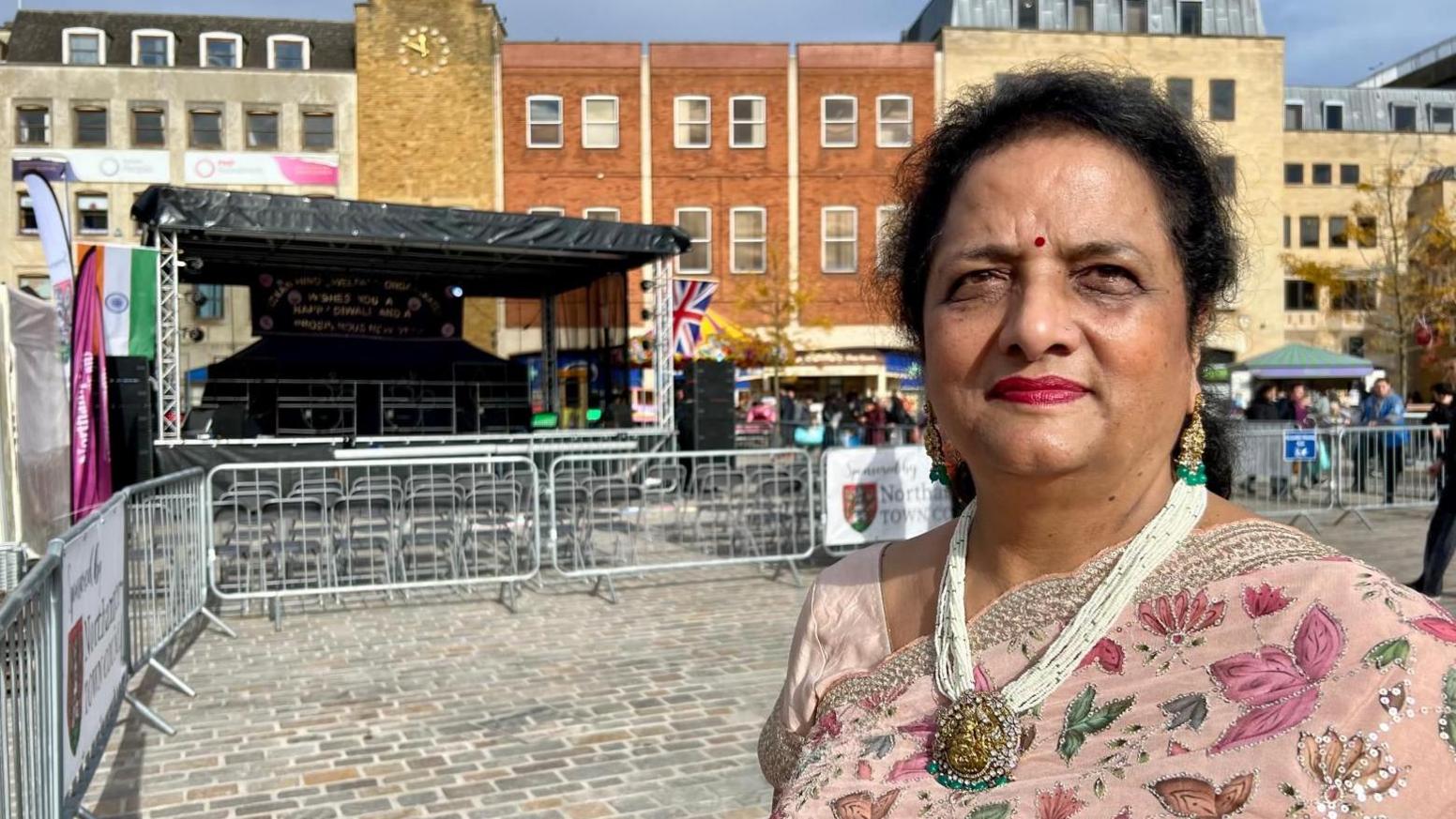 Neelam Aggarwal-Singh, who has tied back black hair and is wearing a pale pink sari embroidered with darker pink flowers and green leaves, edged with sequins over a pink silk top. Over the garments is a chunky necklace. She is standing in Northampton's Market Square and behind her can be seen barriers, a covered raised stand and beyond that buildings.