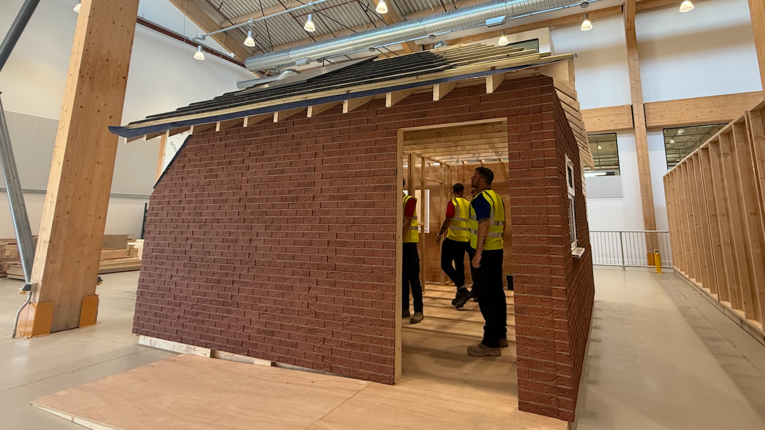 A replica of a partially-built new build home inside the HICSA. It has bricked walls on the exterior, and grey slates on its roof. Inside the building, several pupils wearing yellow high visibility jackets are inspecting it.