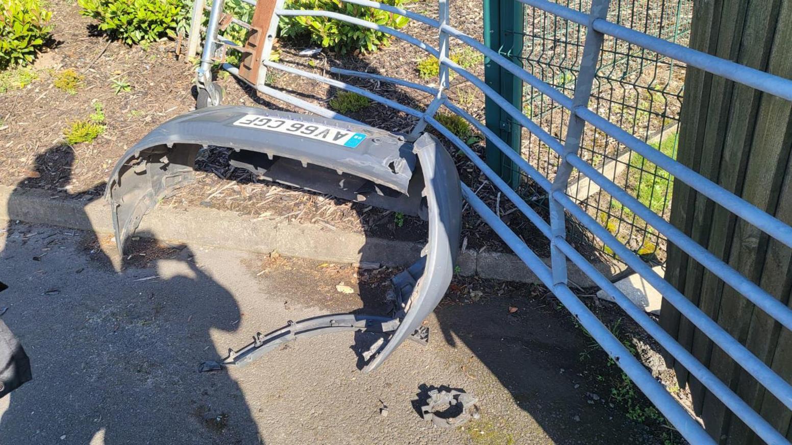 A metal gate which is heavily dented, with the bumper of a van in front of it, including the number plate, and the shadows of two people visible on the ground