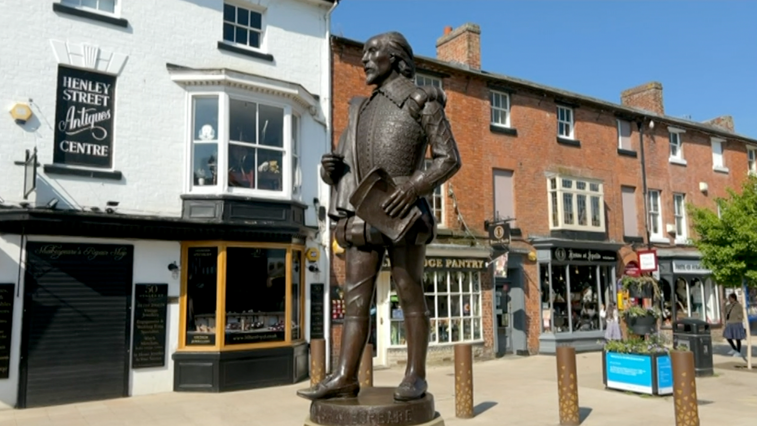 A statue of Shakespeare standing in Stratford-upon-Avon town centre. It is brown and he is holding sheets of paper in the square. 