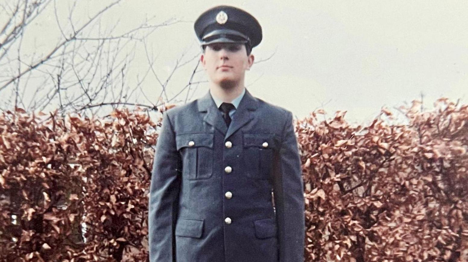 Simon Goodge is dressed in a RAF uniform with gold buttons. He is stood in front of a hedge which is orange in the autumn. The sky is grey