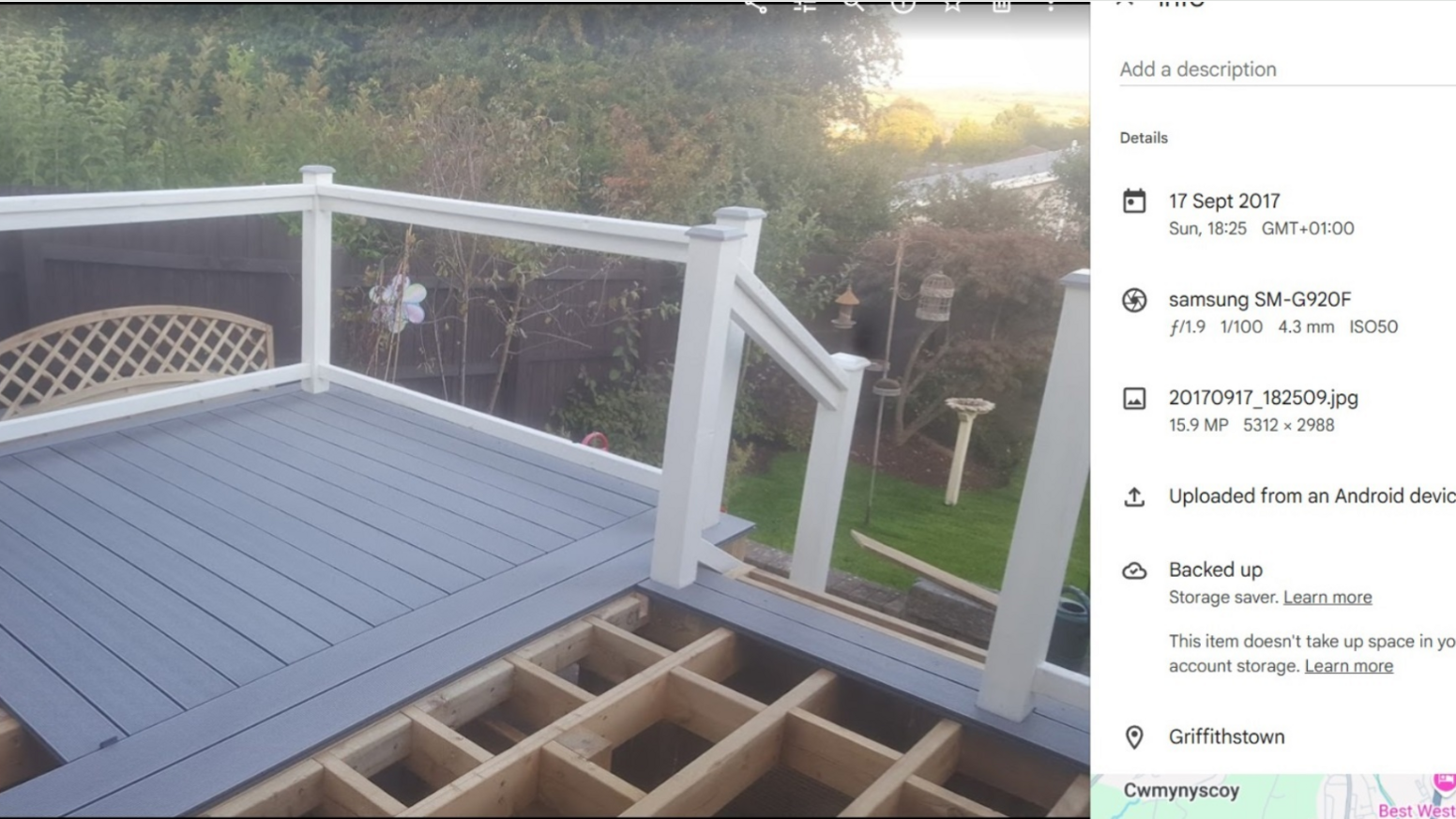 Grey decking with white fencing can be seen raised from a small garden. At the forefront of the image, a raised wooden platform can be seen where the decking floor is unfinished. On the right hand side, the location and time stamp of the image can be seen dated to the 17 Sept 2017.