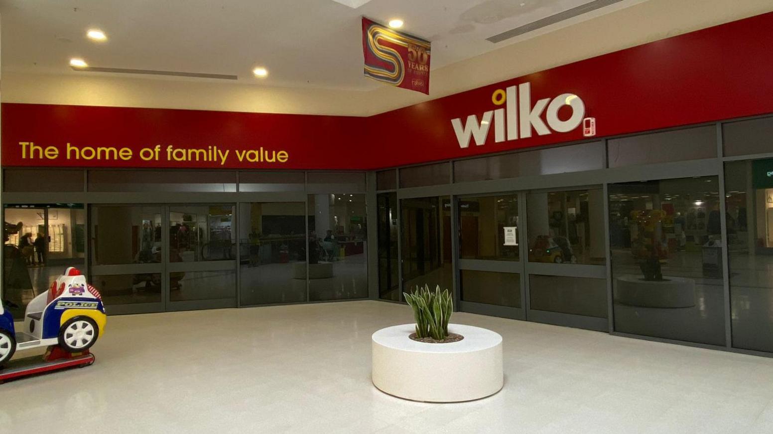 The shop frontage of the old Wilko store in Weston Favel - a black glass front with a red sign and a child's police ride in front and a small decorative plant at the entrance.