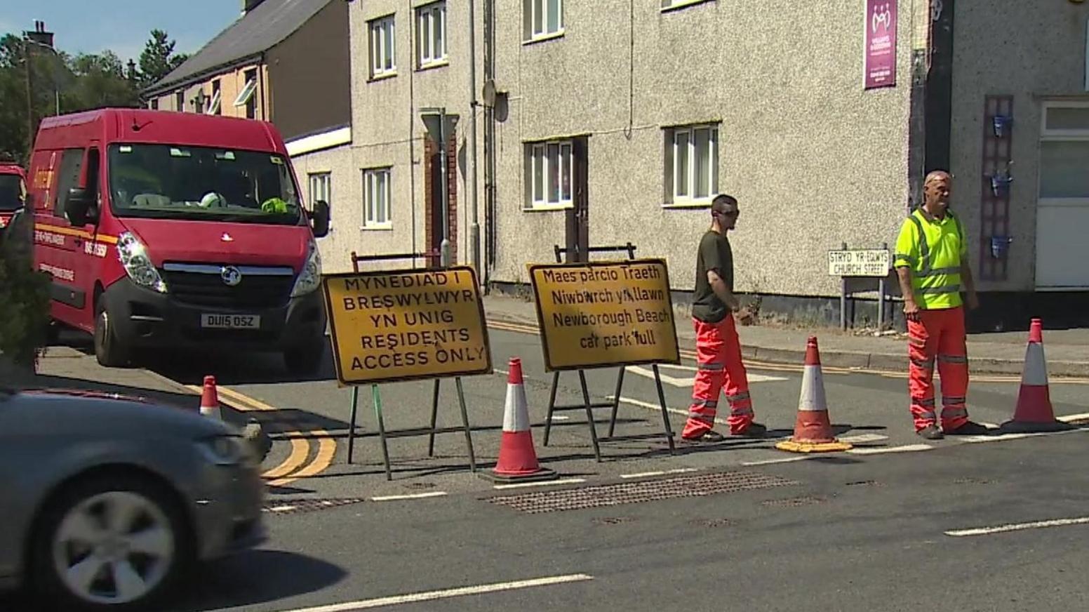 Road in the centre of Newborough with yellow closure signs, cones and traffic management staff