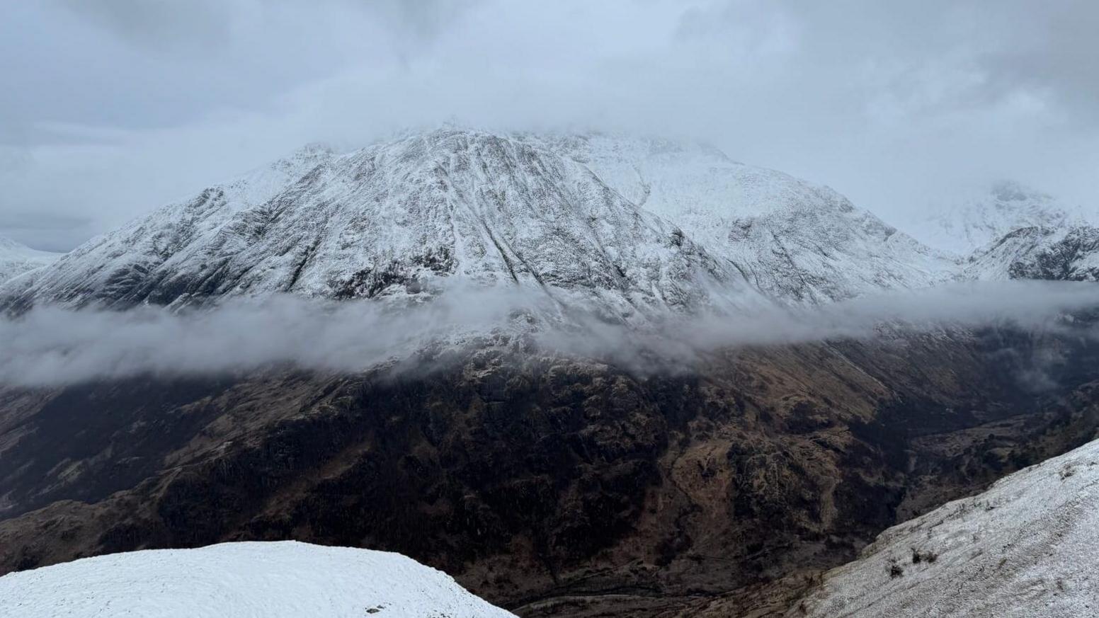 A long line of low cloud lies where a snowline starts on the mountain. 