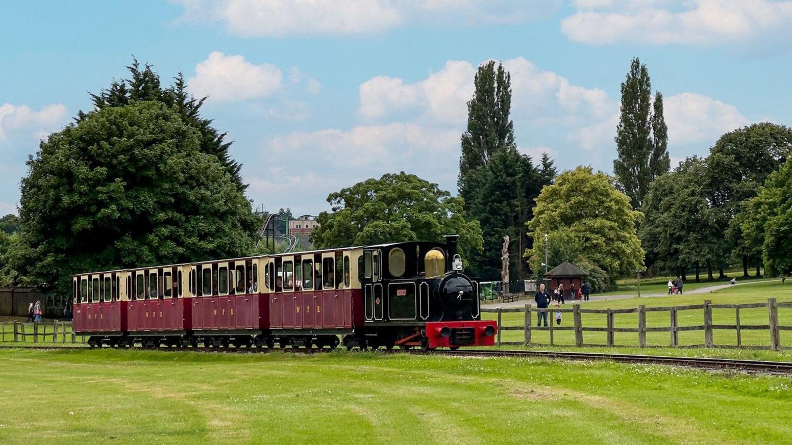 Narrow-gauge railway on grass. A four-carriage train in red and cream livery pulled by a black steam engine.  There is a wooden fence beyond the train and people are seen watching the train.