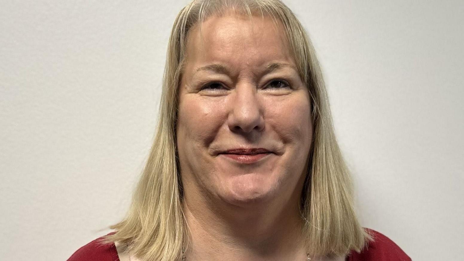 Catherine has shoulder-length blonde hair and is wearing a red top and a long necklace. She is smiling at the camera against a plain white backdrop.