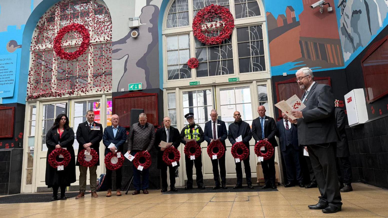 The picture shows a row of people all holding wreaths covered in poppies. They're standing in a train station, and are all looking at a man to the right hand side of them, who is reading an order of service.