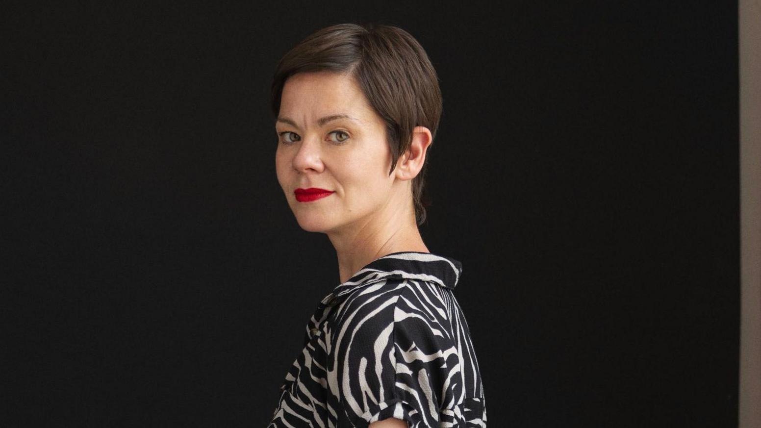 Laura Brewis smiling and  looking over her shoulder. She has a short, straight brown hair and wears a midi length zebra print dress with short sleeves.