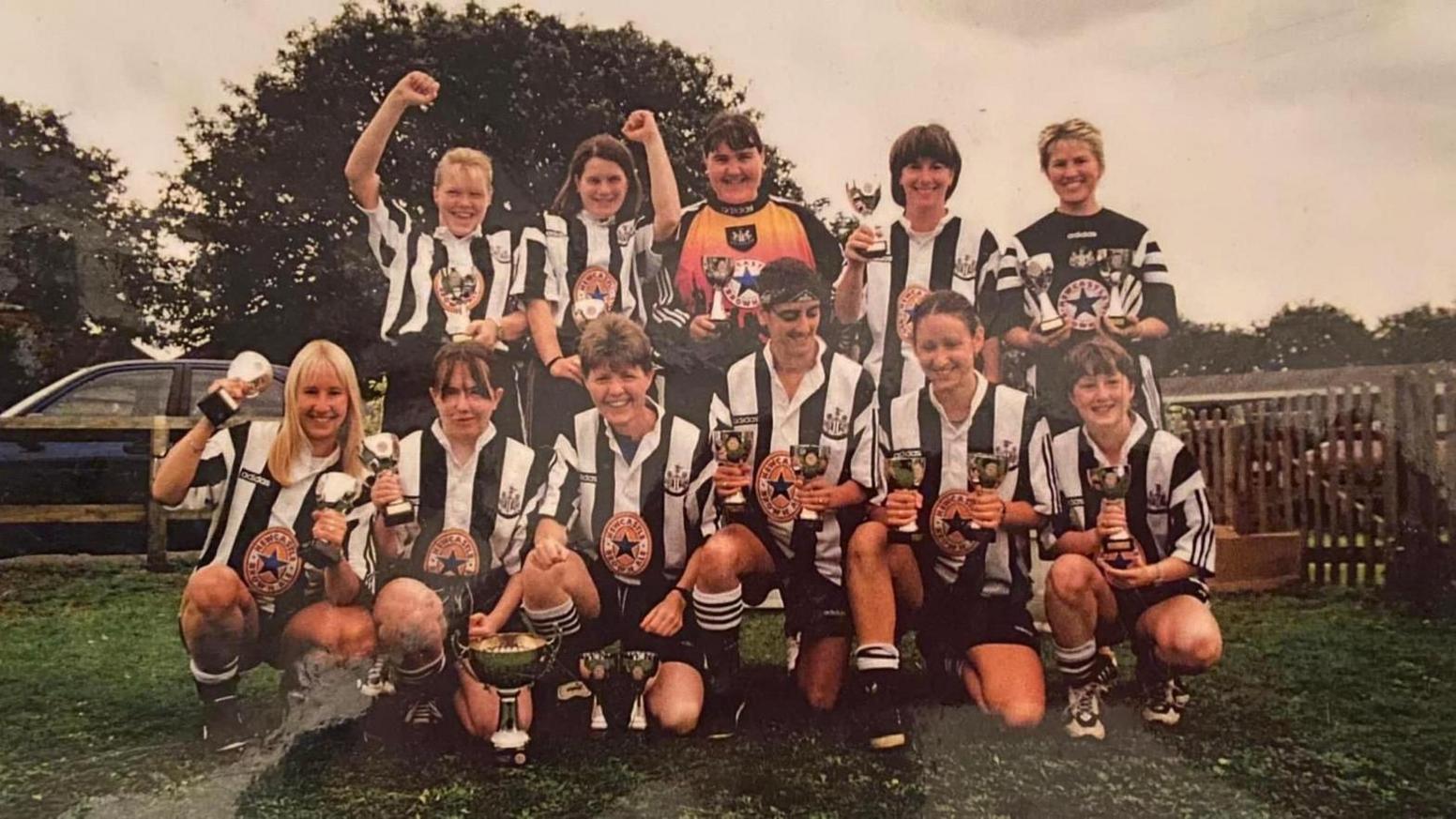 Newcastle Women FC women, wearing black and white striped football shorts, and black shorts and football boots, crouch down and some stand up, holding trophies on a grass field. 