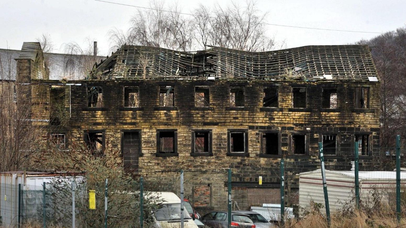 A stone built mill building with empty windows and an exposed roof with collapsed beams