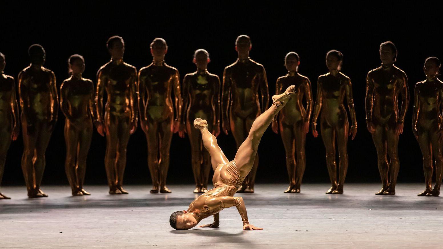 A dancer is balancing on both arms with their head to the floor and legs extended upwards in a diagonal position, showing their strength and flexibility. They are dressed in a shimmering gold costume that reflects the stage lights.
In the background, a line of performers stands evenly spaced, all wearing matching metallic gold bodysuits. 