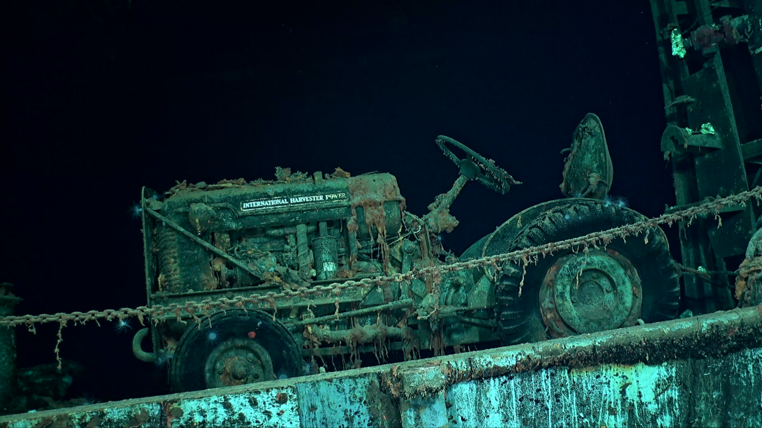 The side view of a tractor on the USS Hornet deck, with a long chain crossing in front of it. The tractor, deck and chain look green, and are covered in bits of seaweed. Behind is the black ocean.