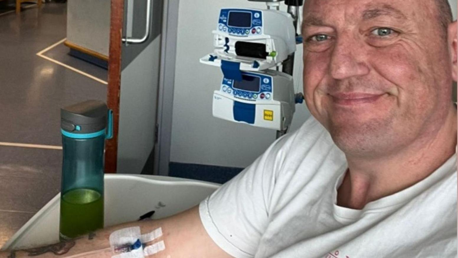 A selfie taken by a man with very short hair wearing a white t-shirt. He is smiling at the camera. Behind him is a small machine and he has an IV line taped onto his arm. Plain walls and floors show he is in a hospital.