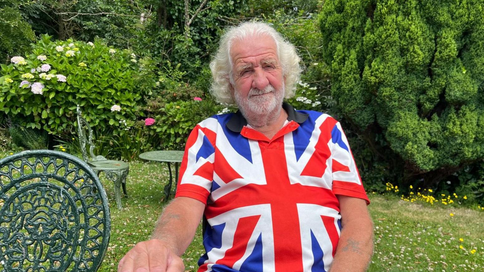 George Le Gallez has white hair and a short white and grey beard and is wearing a union jack shirt in a garden