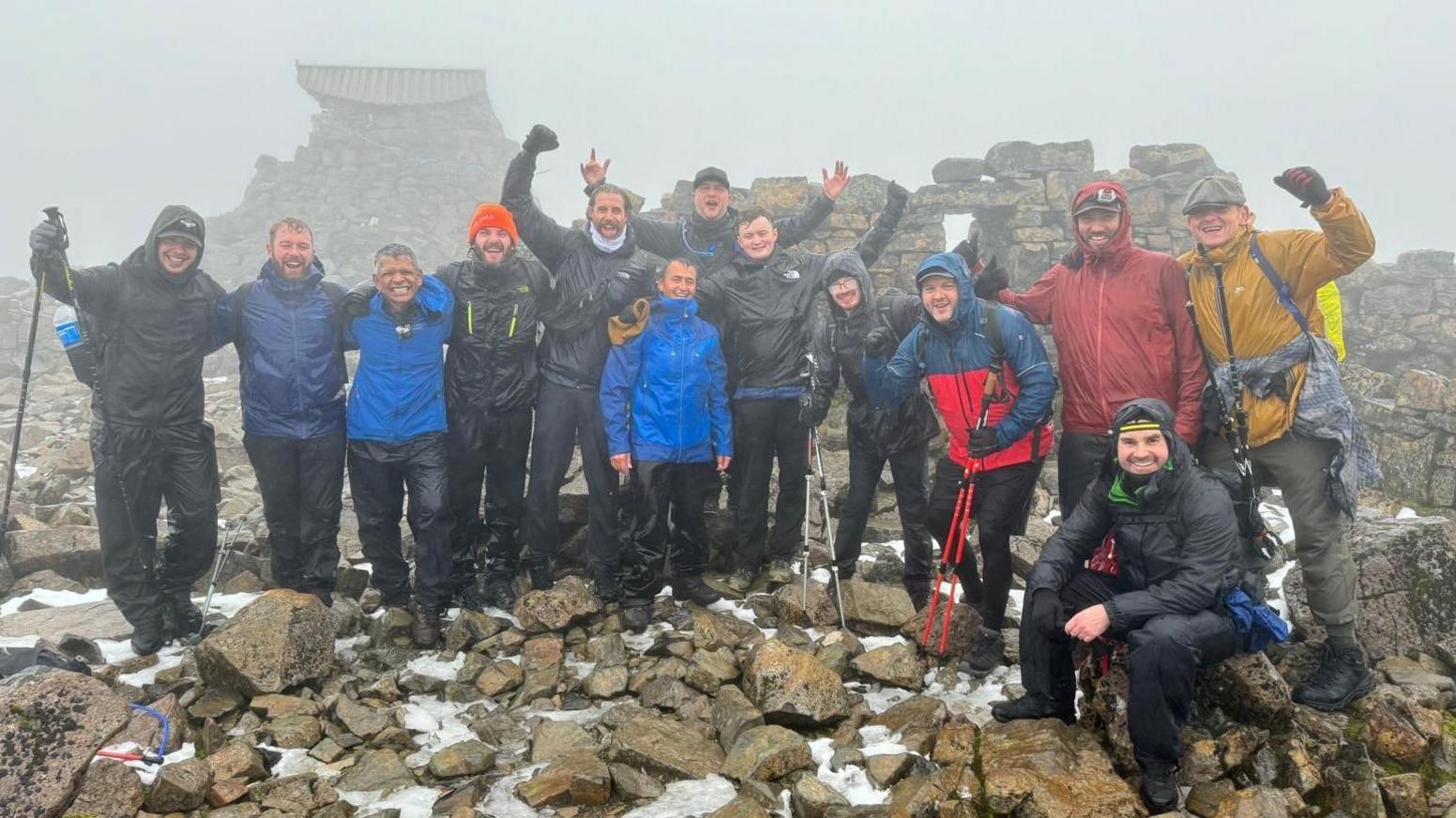 The group at the summit of Ben Nevis. They are celebrating and holding their hands in the air. It is cloudy and wet.