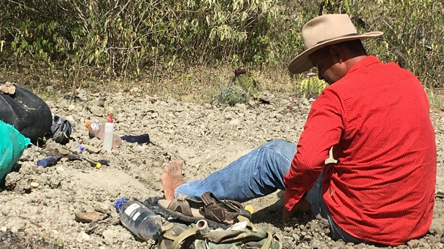 The image shows a fossil collector at a dusty site in Colombia. The man wears a red shirt and a wide-brimmed hat to protect him from the sun. His feet are bare and he is examining the ground carefully for fossils. 