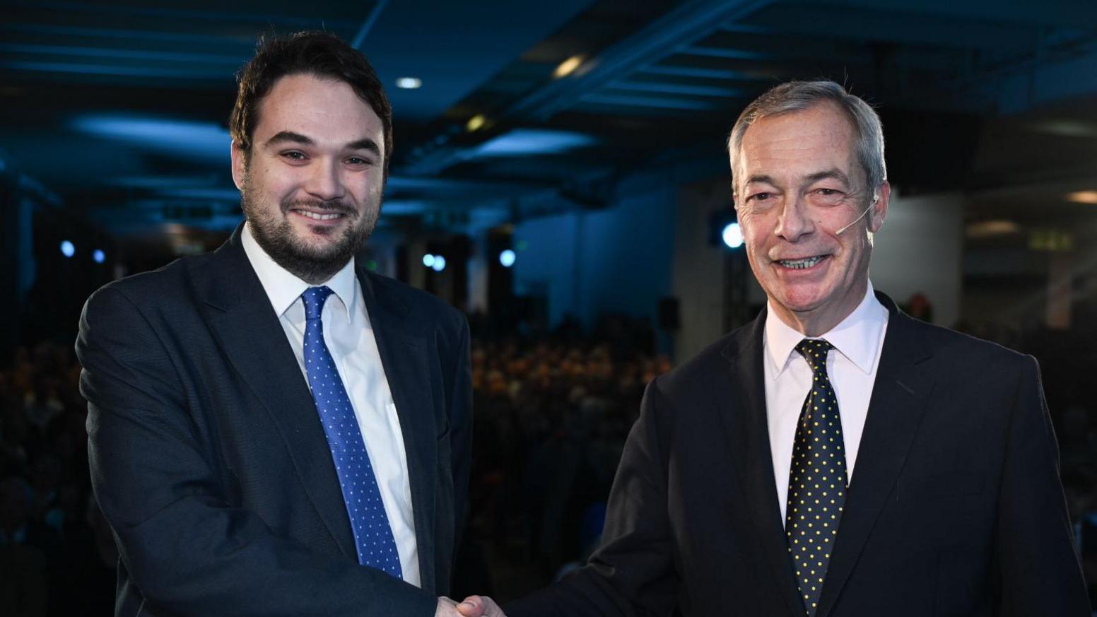 Robin Hunter-Clarke posing for a handshake with Nigel Farage. Both are wearing dark suits. Hunter Clarke has a blue spotted tie, Farage has a black spotted tie.