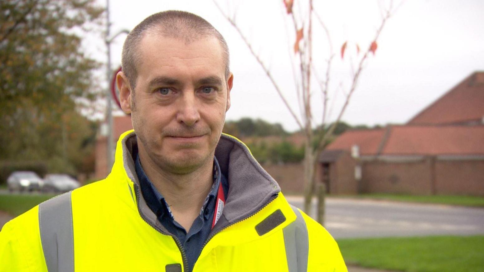 A man with very short hair and blue eyes smiling into the camera and wearing a yellow hi-vis jacket and a shirt. He is standing next to a road with a grass verge either side with a few blurred cars visible behind him.