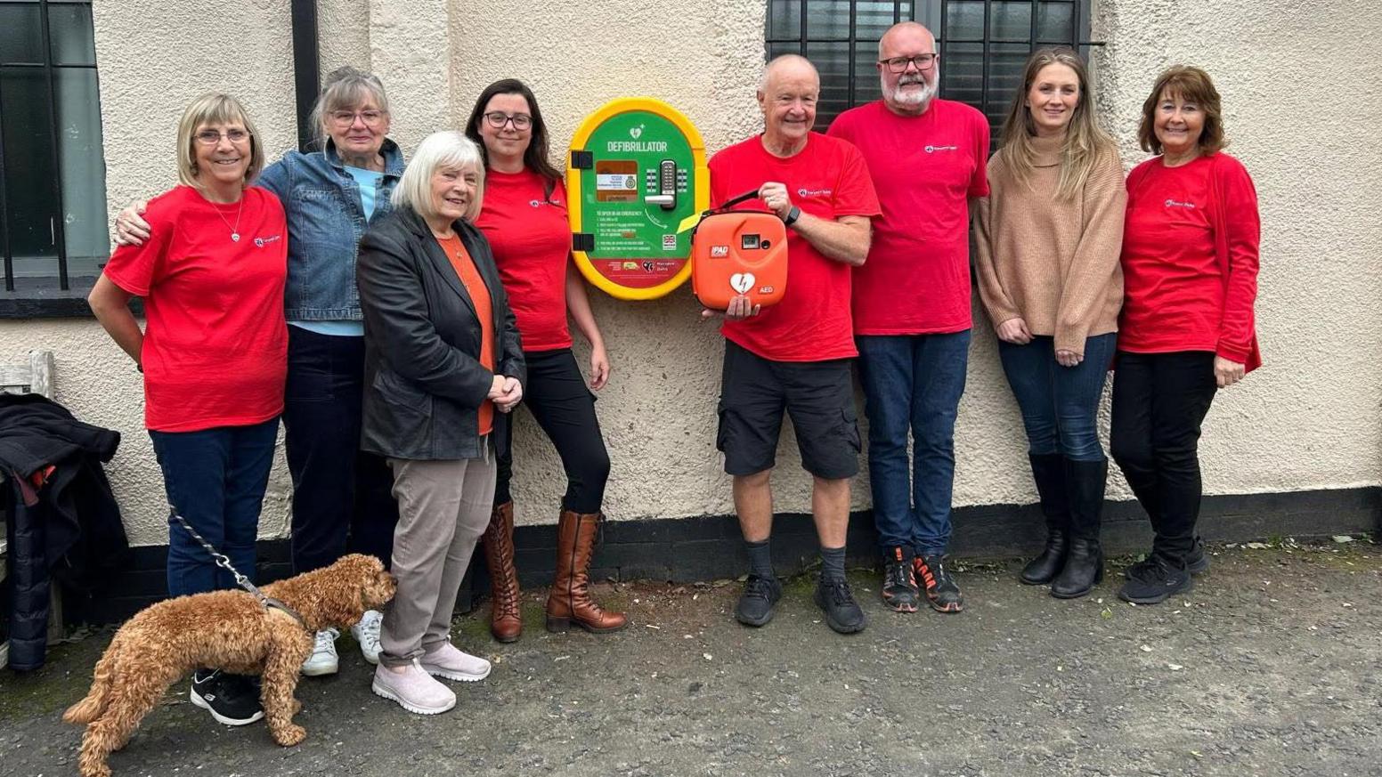 A group of eight people, four wearing red t-shirts and others in casual clothing pose in front of a new defibrillator machine. One of the people in the picture is holding a dog on a leash.