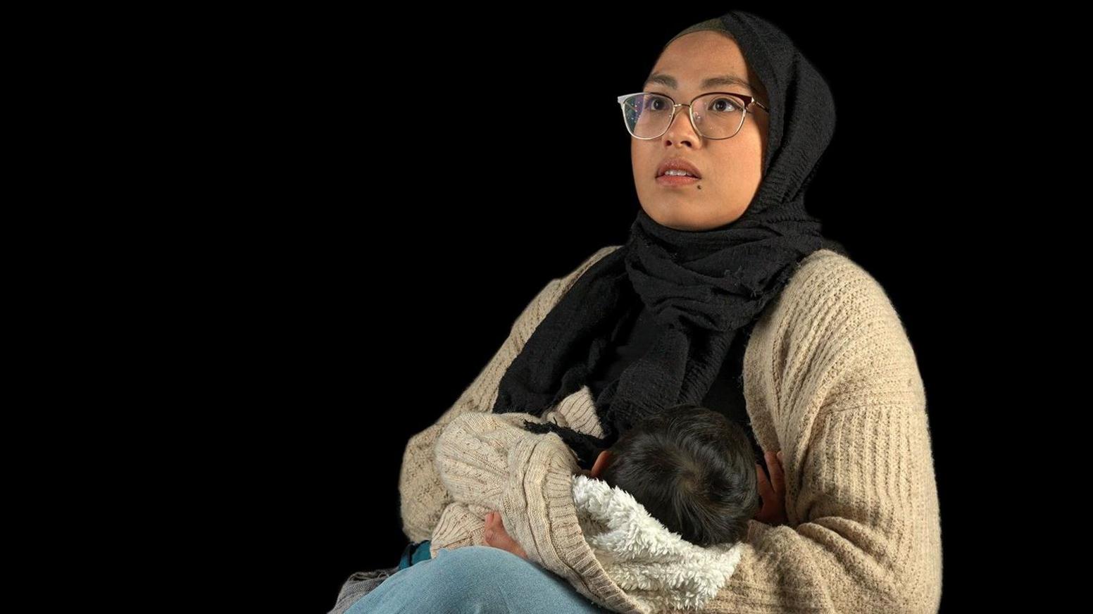 A woman wearing glasses and a headscarf holds a baby in her lap