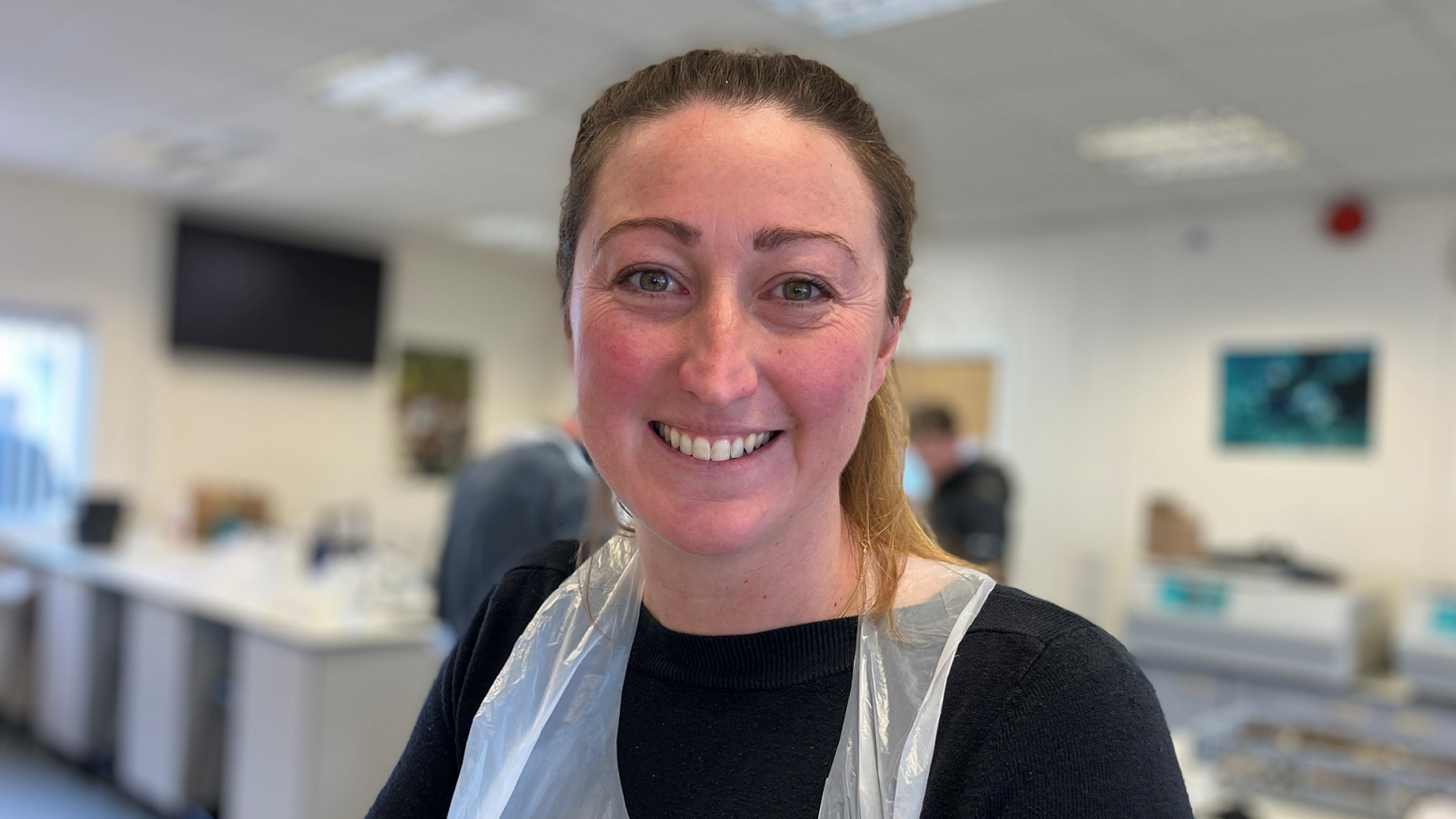 Lottie Johns, dressed in a black jumper and standing in the marine laboratory, is wearing a white plastic apron and is smiling for the camera. She has long brown hair, which has been tied in a ponytail. 