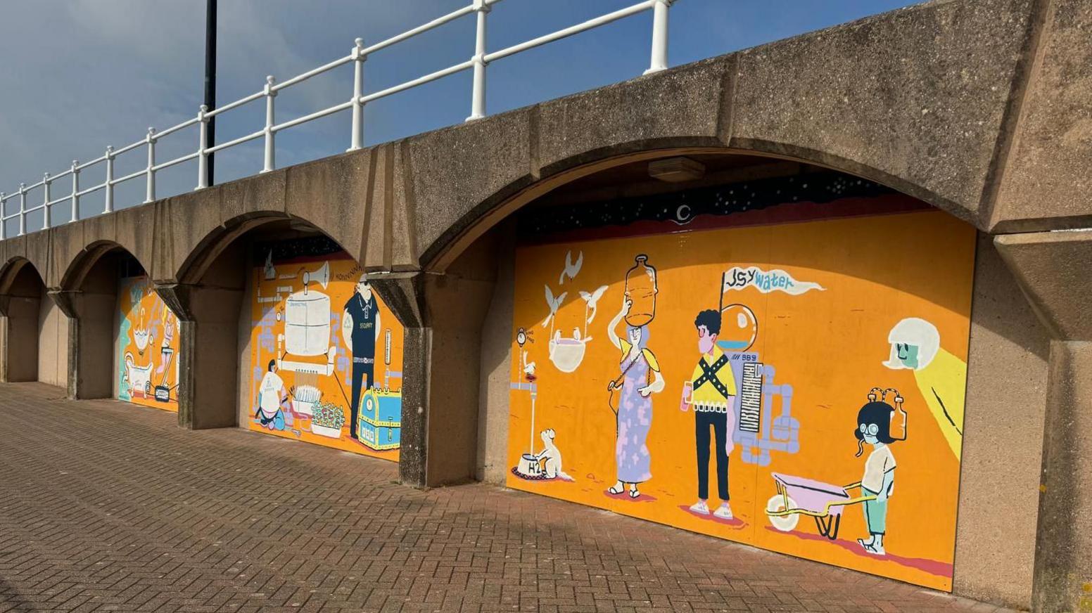 Three murals on the Elizabeth Marina in Jersey, orange backgrounds with cartoon people, railings above, grey and blue skies