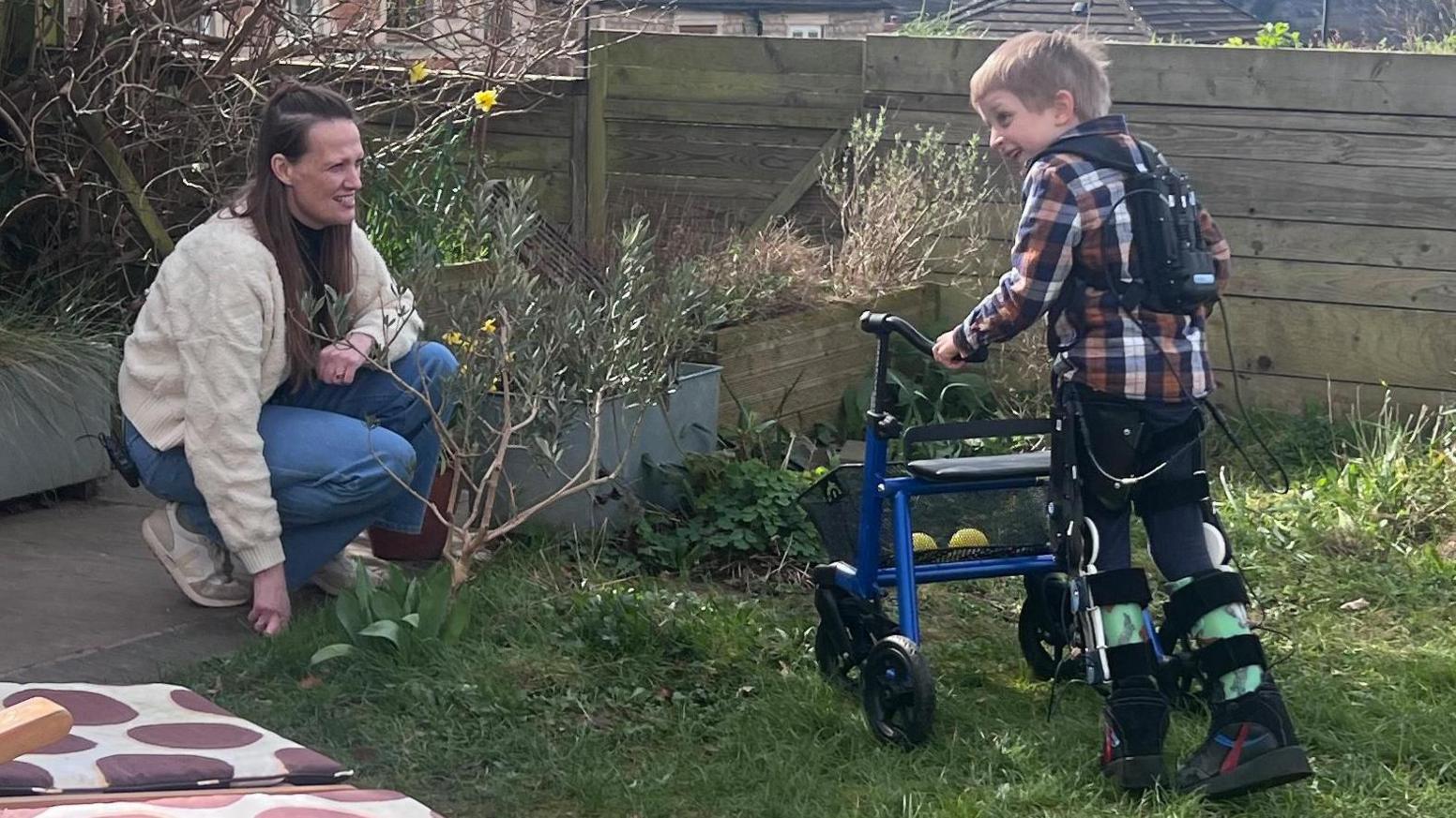 Asger, a young boy with blonde hair, is pushing a wheeled walking aid in front of him, while wearing an exoskeleton. The exoskeleton comprises leg braces and what looks like a power bank worn as a backpack. He is walking towards his mum Holly, who is crouching down to his level, in their garden.