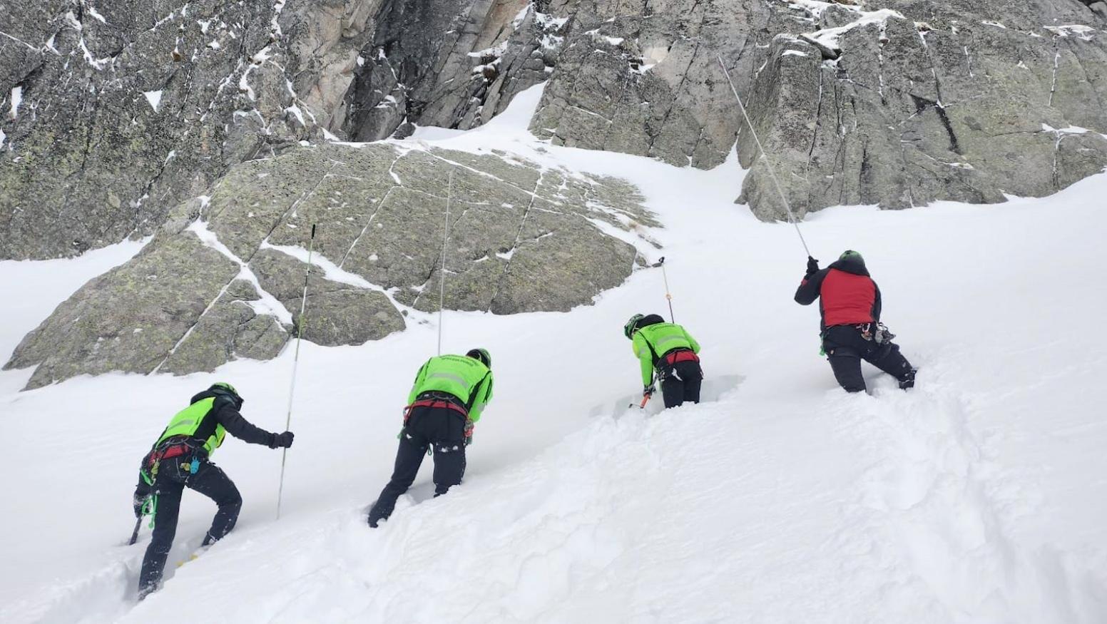 Four alpine rescue search team members wearing high vis uniforms search the snowy side of a mountain with ice picks and long poles. 