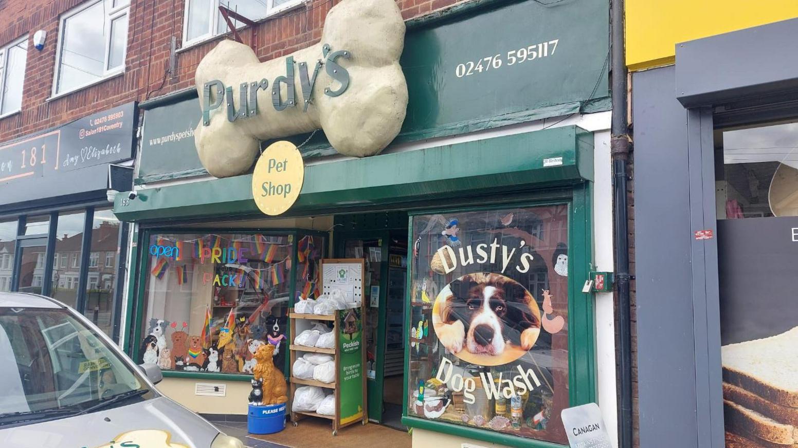 The frontage of a pet food shop. A large sign in the shape of a bone that says Purdy's Pet Shop sits above the door. The window frames are painted green, on the right window  is a picture of a dog in a lying/begging position that says Dusty's Dog Wash.