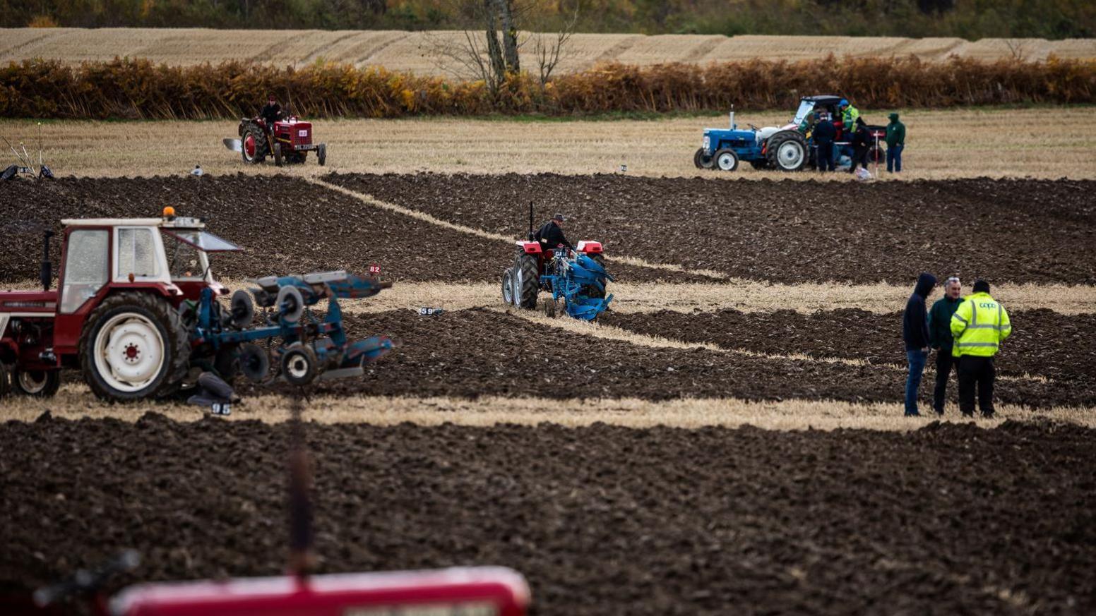 Five tractors of different types ploughing in a field. A group of three people are gathered talking.