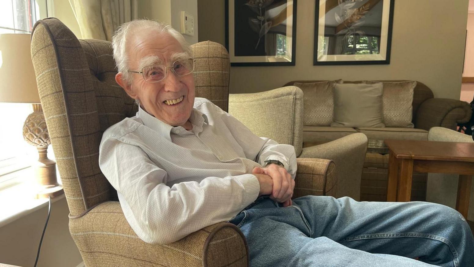 An elderly man sits smiling in a chair in the living area of a nursing home.