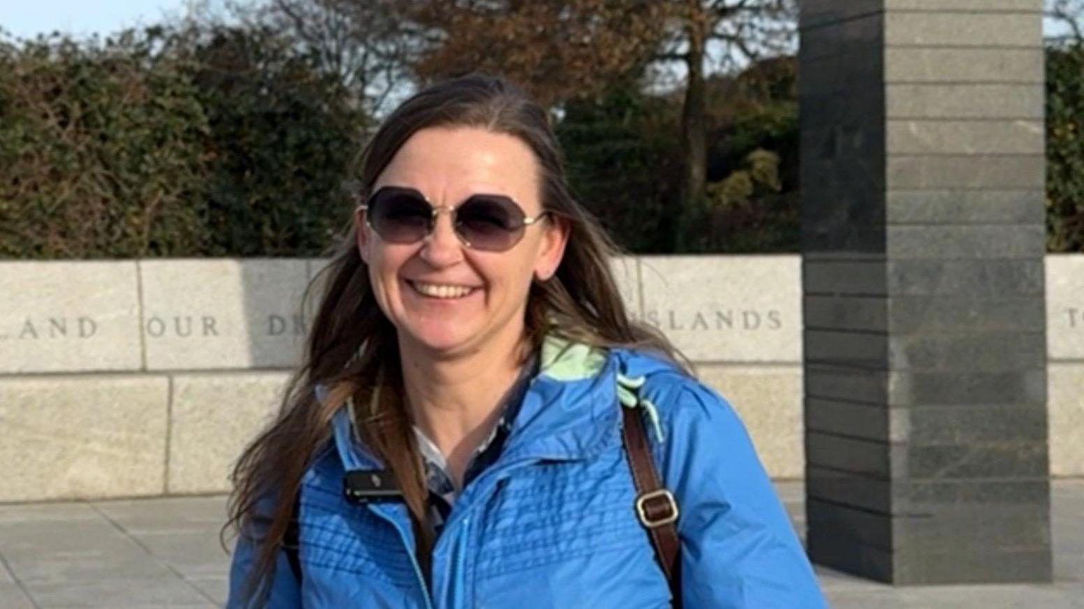 A lady is pictured wearing a blue coat and a black bag. She has brown hair and is wearing glasses. She is smiling.