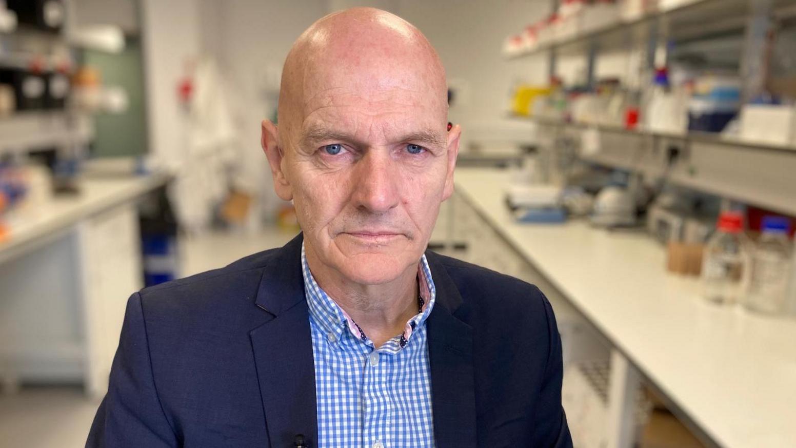 Prof Chris Elliott is wearing a blue gingham shirt and a blue blazer. He is standing in a laboratory and looks sternly towards the camera