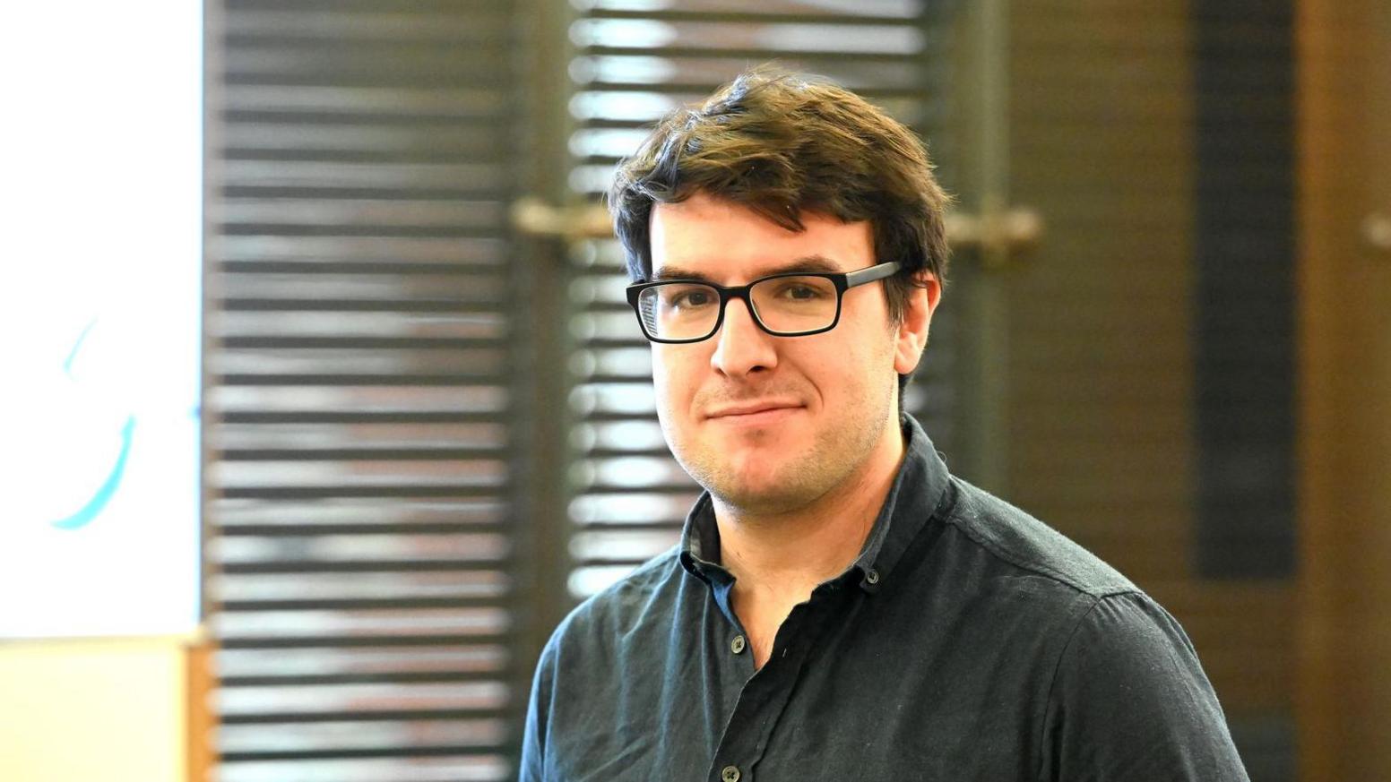 A portrait of a man with dark hair and glasses wearing a dark shirt. He is standing in front of venetian blinds. 