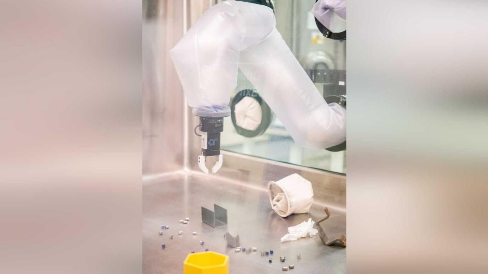 A robotic arm inside an active nuclear glovebox. The clear box has a a robotic arm with a flexible arm inside it. There are some metal objects inside the box, as well as a yellow hexagonal box.