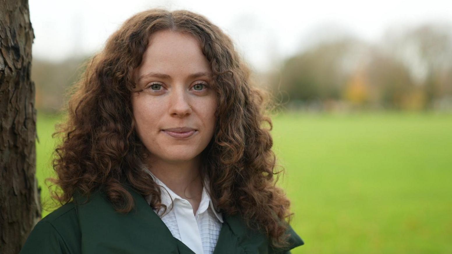 Holly stares to camera. She is in focus with dark, long curly hair and is leaning against a tree-trunk while the background is out of focus but looks like a green field.