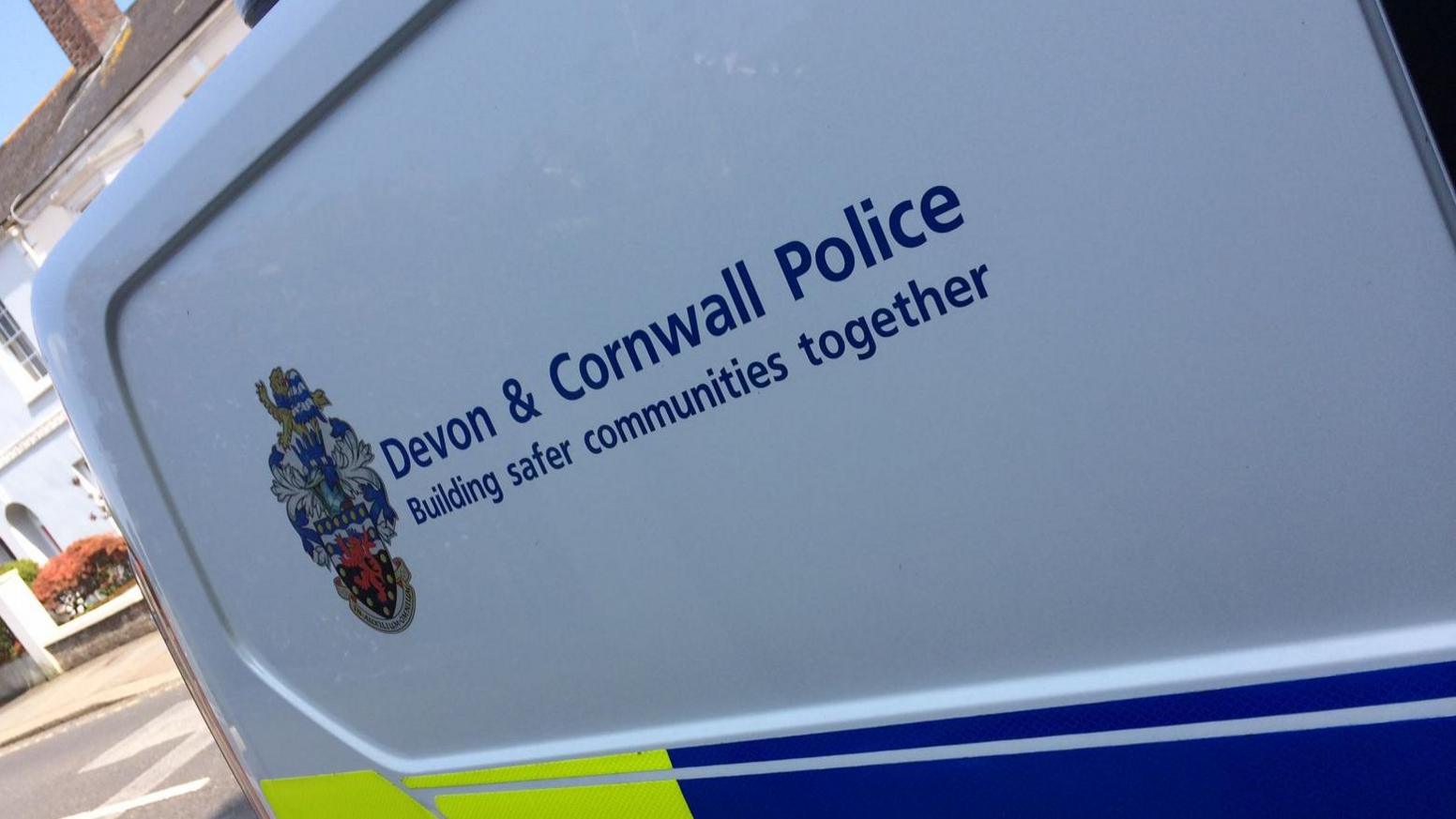The side of a police van. It is blue and green in colour with a white gap - where the Devon and Cornwall Police logo is. Below the logo and text it says - 'Building safer communities together'