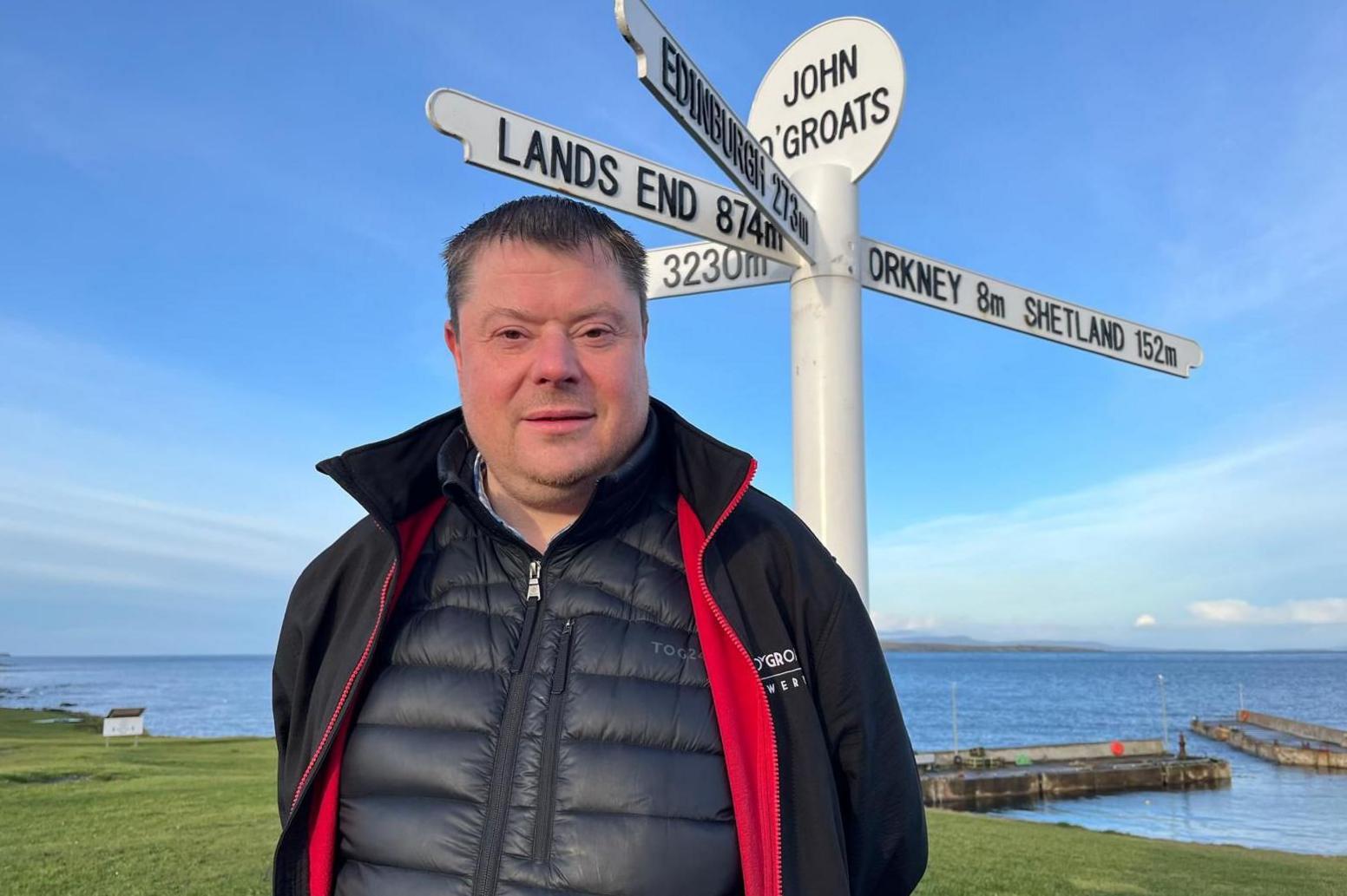 Andrew is standing next to a sign for John O'Groats. The sign points to other places including Lands End, Edinburgh and Orkney and Shetland.