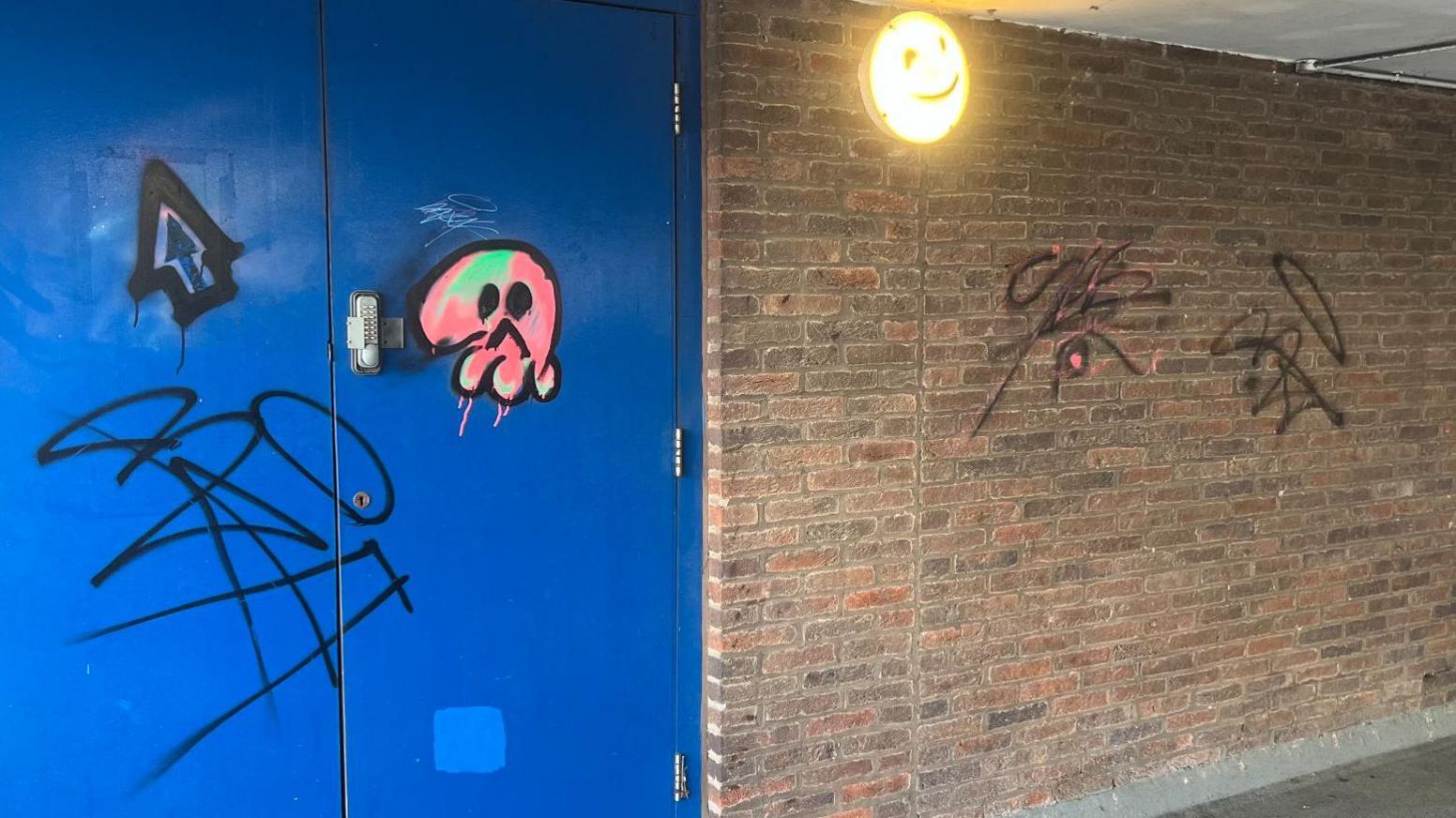 A blue double door has three pieces of graffiti on it - one is an arrow, one is a general scrawl/tag and the third is a pink, alien-type design with two eyes and a ^ as a mouth. A red-brick wall next to the door also has two graffiti tags on it which.