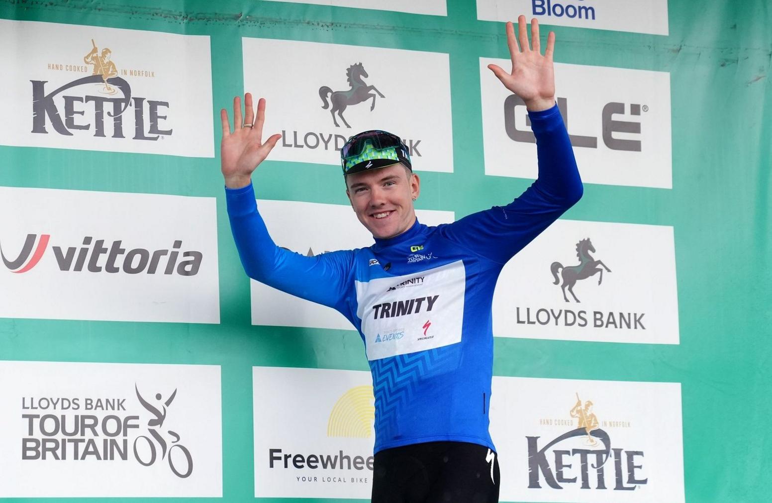 A man wearing blue lyra top and sunglasses on his head holds his hands high and smiles as he stands on the podium