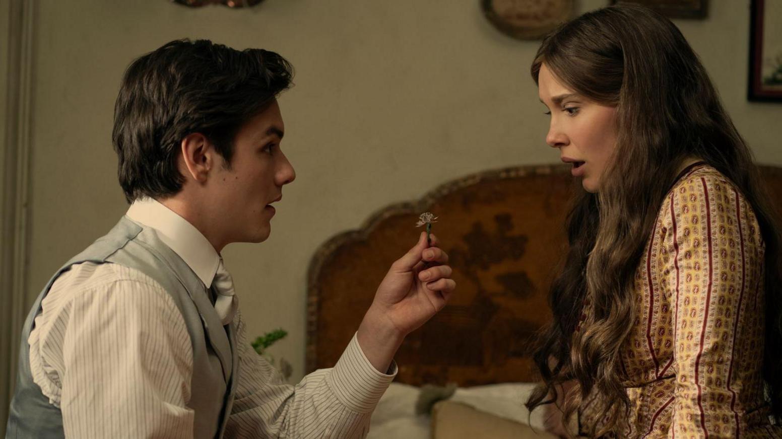 Louis Partridge and Millie Bobby Brown in Enola Holmes. He is holding a ring and she is sitting on the bed looking surprised