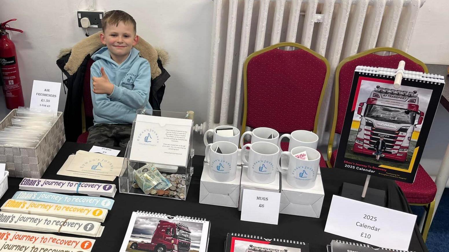 A boy in a turquoise hoodie with his thumb raised, sitting on a chair in front of a stall of merchandise printed with the Ethan's journey to recovery slogan. Stickers, mugs and a 2025 calendar can be seen.