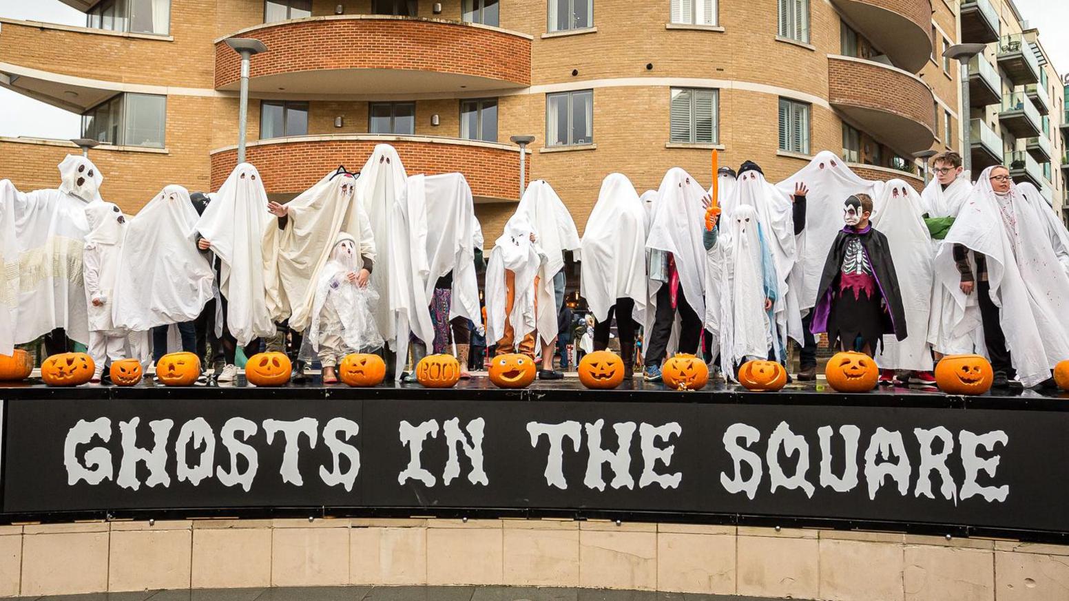 A group of people with white bed sheets over them - appearing like ghosts. They are stood atop a sign saying 'Ghosts in the Square'.